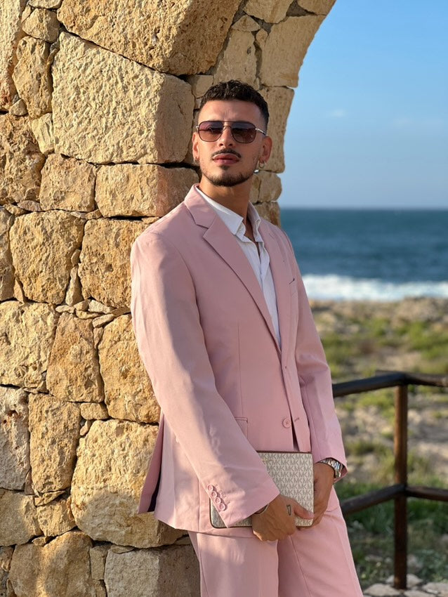 Mann in rosa Anzug steht vor einer Steinmauer mit Blick auf das Meer
