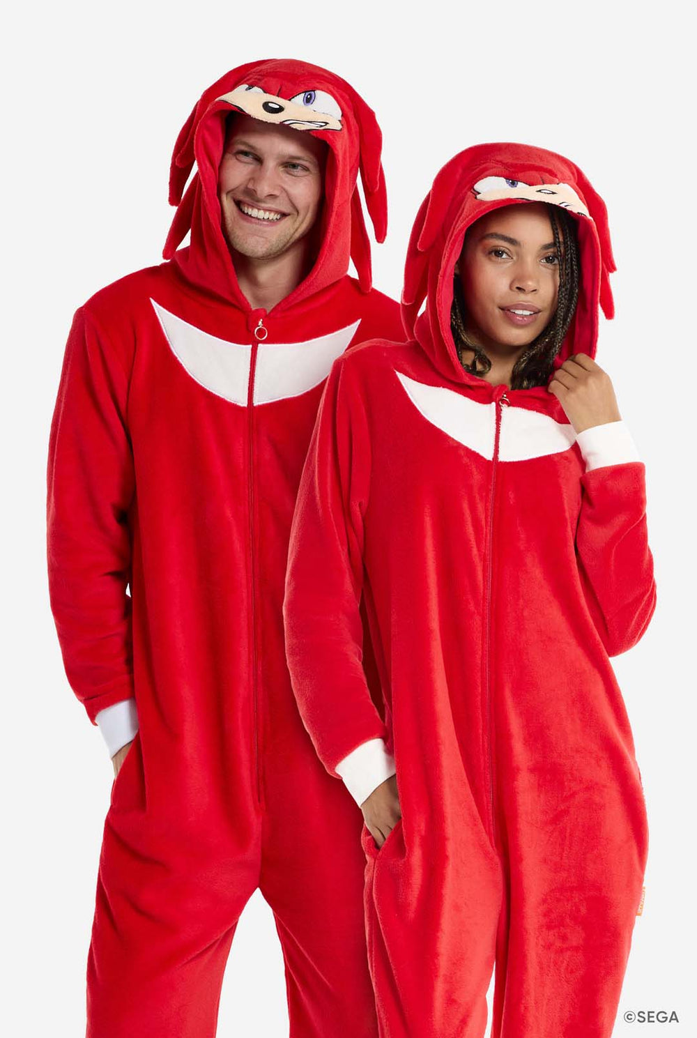 Ein Mann und eine Frau tragen einen knallroten Knuckles the Echidna-Strampler für Erwachsene von OppoSuits. Der lustige und kuschelige Strampler hat einen Reißverschluss vorne und eine Kapuze, die Knuckles' Gesicht nachempfunden ist, mit ausdrucksstarken Augen und schlaffen Dreadlocks. Der Anzug ist hauptsächlich rot, mit einem weißen Halbmondmuster auf der Brust, das Knuckles' ikonischen Look aus der Sonic the Hedgehog-Videospielserie widerspiegelt.