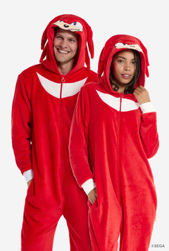 Ein Mann und eine Frau tragen einen knallroten Knuckles the Echidna-Strampler für Erwachsene von OppoSuits. Der lustige und kuschelige Strampler hat einen Reißverschluss vorne und eine Kapuze, die Knuckles' Gesicht nachempfunden ist, mit ausdrucksstarken Augen und schlaffen Dreadlocks. Der Anzug ist hauptsächlich rot, mit einem weißen Halbmondmuster auf der Brust, das Knuckles' ikonischen Look aus der Sonic the Hedgehog-Videospielserie widerspiegelt.