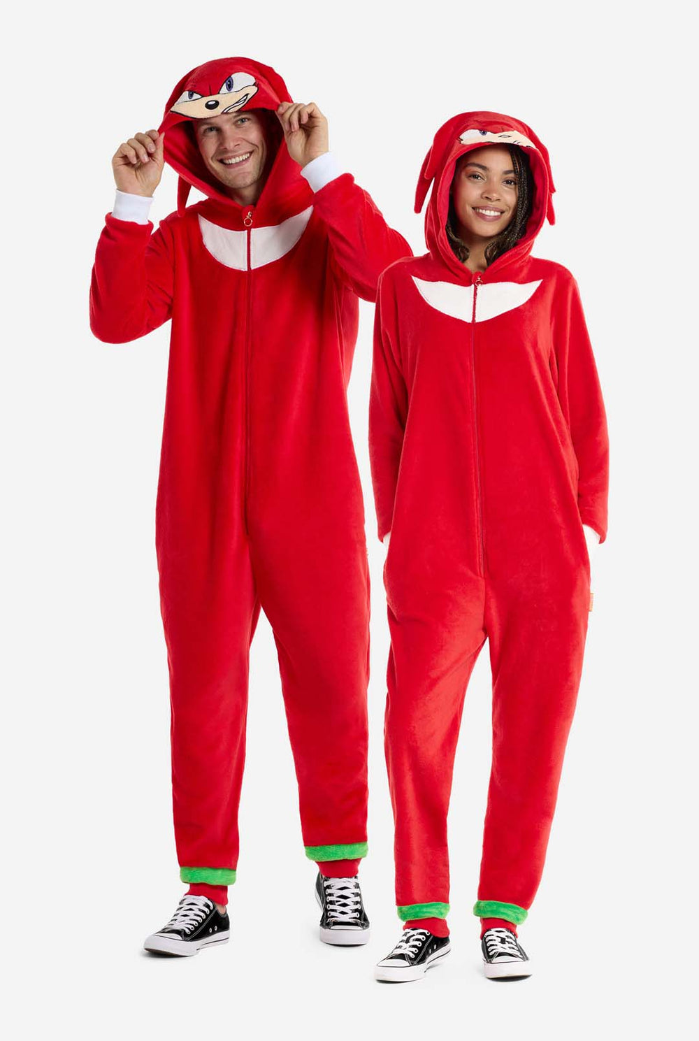 Ein Mann und eine Frau tragen einen knallroten Knuckles the Echidna-Strampler für Erwachsene von OppoSuits. Der lustige und kuschelige Strampler hat einen Reißverschluss vorne und eine Kapuze, die Knuckles' Gesicht nachempfunden ist, mit ausdrucksstarken Augen und schlaffen Dreadlocks. Der Anzug ist hauptsächlich rot, mit einem weißen Halbmondmuster auf der Brust, das Knuckles' ikonischen Look aus der Sonic the Hedgehog-Videospielserie widerspiegelt.