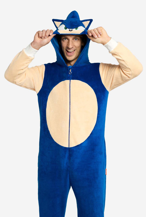 Ein Mann trägt einen verspielten blau-beigen Sonic the Hedgehog-Onesie für Erwachsene von OppoSuits. Der kuschelige Onesie hat einen Reißverschluss vorne und eine angenähte Kapuze, die Sonics ikonisches Gesicht mit Ohren und ausdrucksstarkem Blick nachempfunden ist. Der blaue Körper des Anzugs kontrastiert mit dem beigen Bauch und imitiert Sonics klassischen Look. Dieser lustige, bequeme und stylische Onesie eignet sich perfekt zum Entspannen, für Mottopartys oder Cosplay-Events.