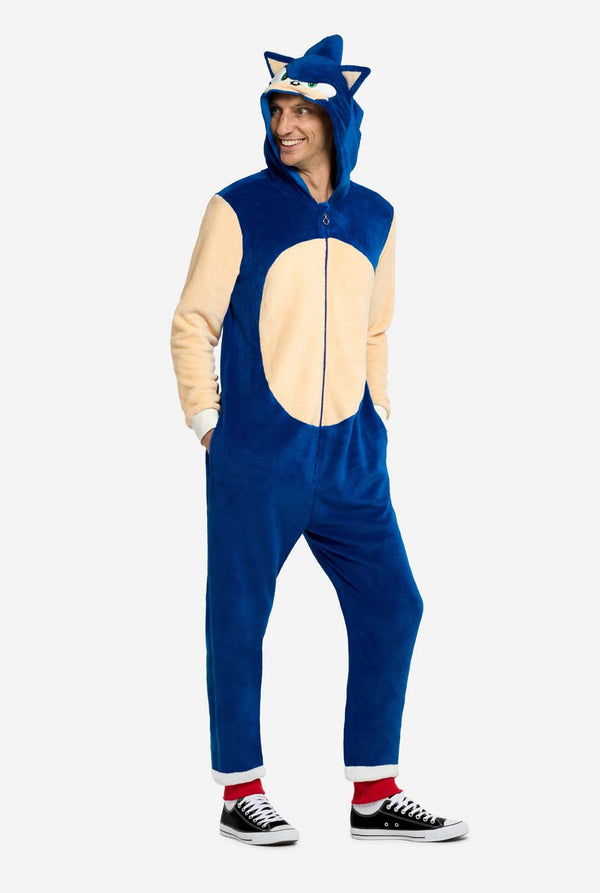 Ein Mann trägt einen verspielten blau-beigen Sonic the Hedgehog-Onesie für Erwachsene von OppoSuits. Der kuschelige Onesie hat einen Reißverschluss vorne und eine angenähte Kapuze, die Sonics ikonisches Gesicht mit Ohren und ausdrucksstarkem Blick nachempfunden ist. Der blaue Körper des Anzugs kontrastiert mit dem beigen Bauch und imitiert Sonics klassischen Look. Er hält seine Hände in den Taschen.