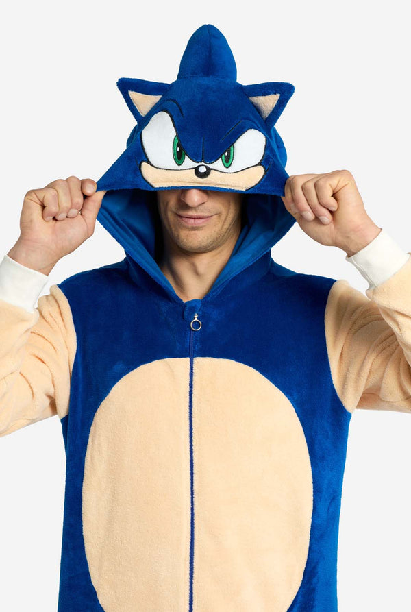 Ein Mann trägt einen verspielten blau-beigen Sonic the Hedgehog-Onesie für Erwachsene von OppoSuits. Der kuschelige Onesie hat einen Reißverschluss vorne und eine angenähte Kapuze, die Sonics ikonisches Gesicht mit Ohren und ausdrucksstarkem Blick nachempfunden ist. Der blaue Körper des Anzugs kontrastiert mit dem beigen Bauch und imitiert Sonics klassischen Look. Er hält sich die Kapuze über die Augen.