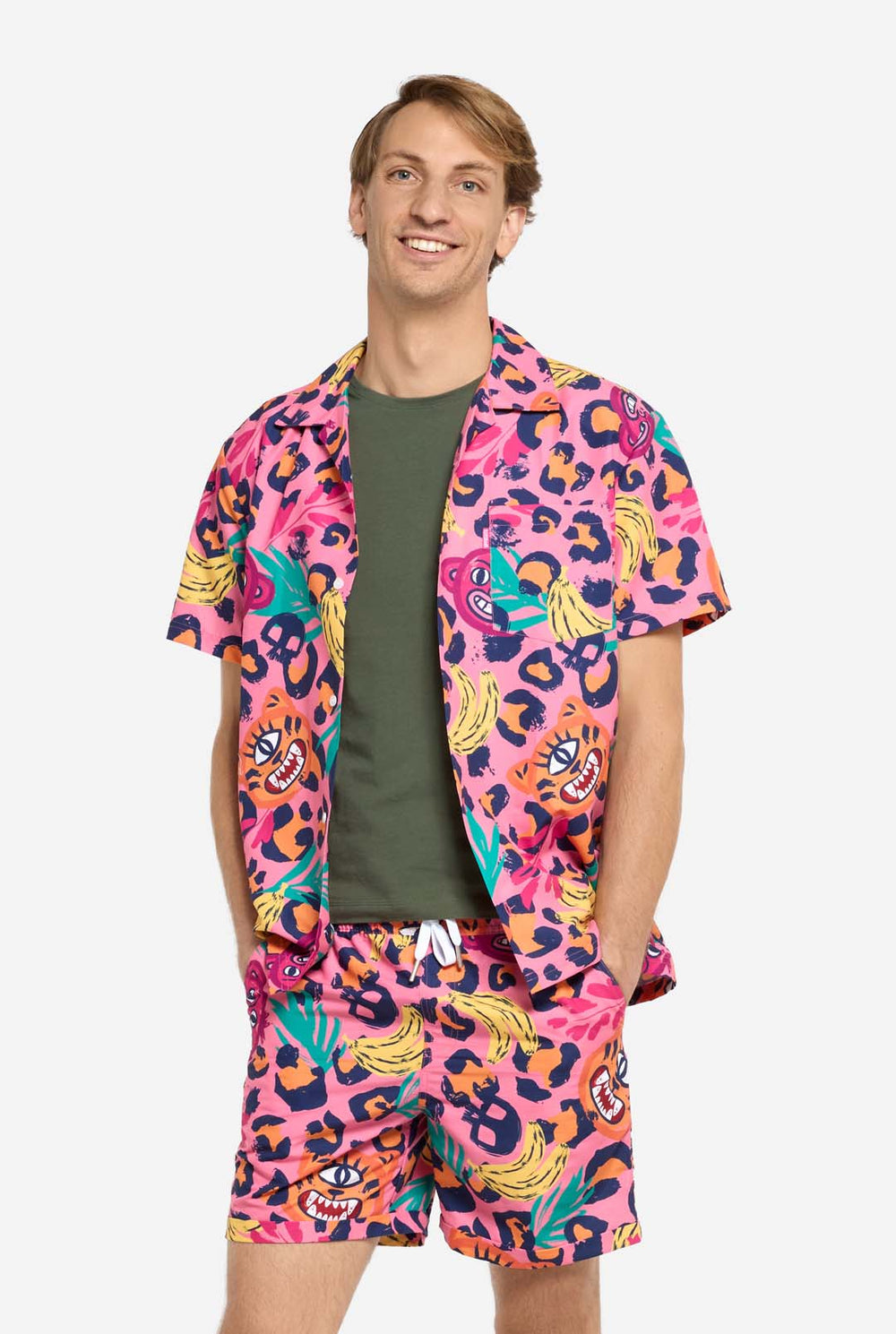 Ein Mann trägt ein farbenfrohes zweiteiliges Sommeroutfit mit auffälligem Tropenmuster. Das kurzärmelige Hemd und die dazu passenden Shorts präsentieren ein lebendiges Design mit neonpinken, orangefarbenen und gelben Leopardenmustern, Bananen, grünen Palmblättern und witzigen Cartoon-Gesichtern. Das Outfit ist lässig mit einem grünen T-Shirt darunter gestylt und perfekt für Sommerpartys, Urlaube oder Spaß am Pool.