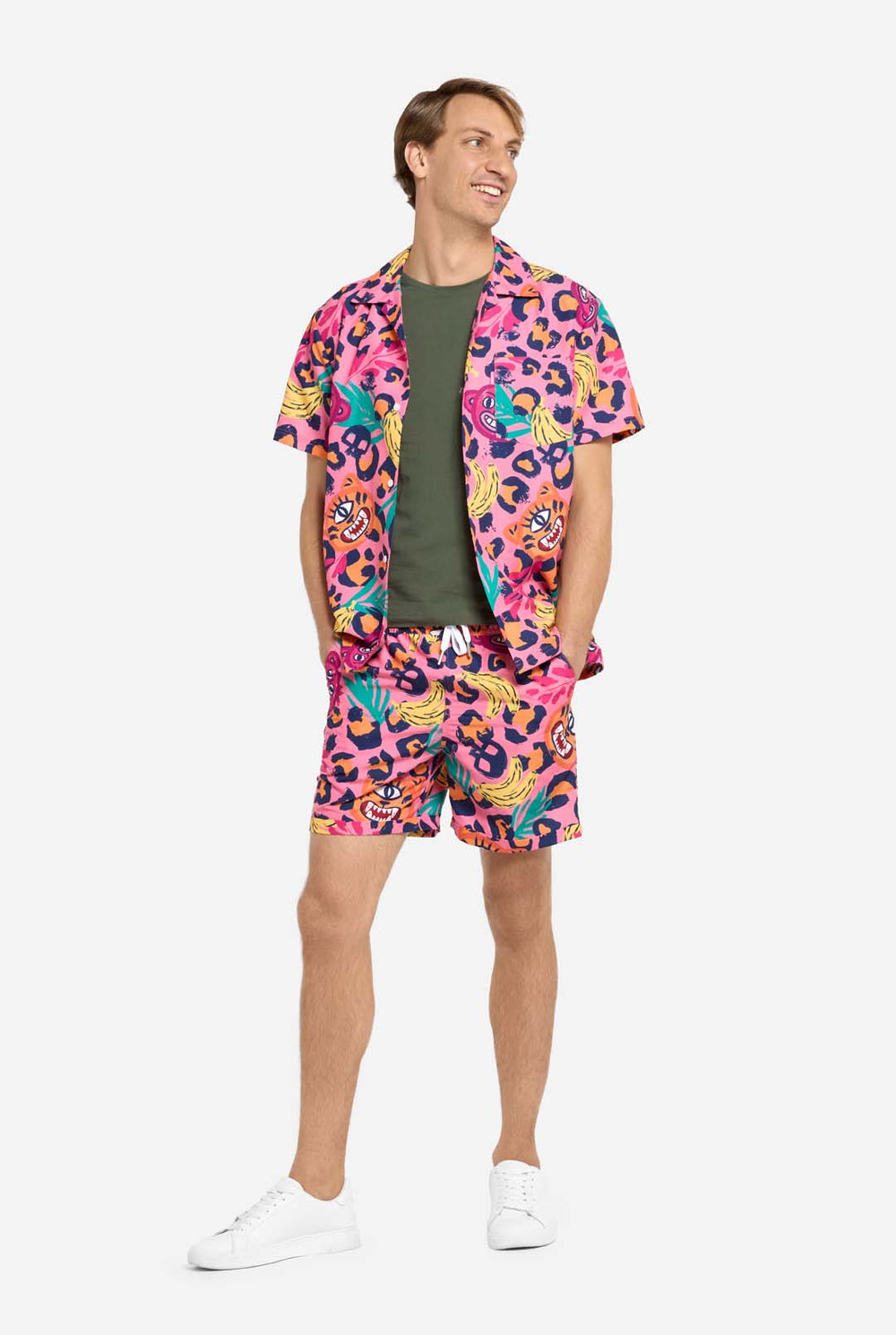Ein Mann trägt ein farbenfrohes zweiteiliges Sommeroutfit mit auffälligem Tropenmuster. Das kurzärmelige Hemd und die dazu passenden Shorts präsentieren ein lebendiges Design mit neonpinken, orangefarbenen und gelben Leopardenmustern, Bananen, grünen Palmblättern und witzigen Cartoon-Gesichtern. Das Outfit ist lässig mit einem grünen T-Shirt darunter gestylt und perfekt für Sommerpartys, Urlaube oder Spaß am Pool.