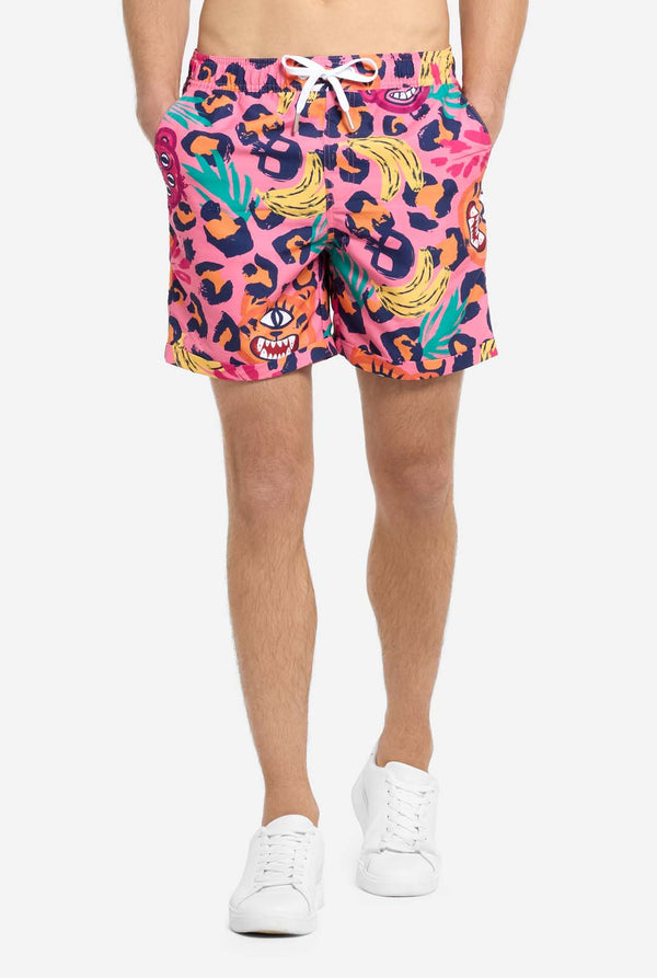 Ein Mann trägt ein farbenfrohes zweiteiliges Sommeroutfit mit auffälligem Tropenmuster. Das kurzärmelige Hemd und die dazu passenden Shorts präsentieren ein lebendiges Design mit neonpinken, orangefarbenen und gelben Leopardenmustern, Bananen, grünen Palmblättern und witzigen Cartoon-Gesichtern. Das Outfit ist lässig mit einem grünen T-Shirt darunter gestylt und perfekt für Sommerpartys, Urlaube oder Spaß am Pool. Nahaufnahme der Shorts.
