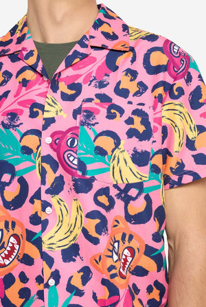 Ein Mann trägt ein farbenfrohes zweiteiliges Sommeroutfit mit auffälligem Tropenmuster. Das kurzärmelige Hemd und die dazu passenden Shorts präsentieren ein lebendiges Design mit neonpinken, orangefarbenen und gelben Leopardenmustern, Bananen, grünen Palmblättern und skurrilen Cartoon-Gesichtern. Das Outfit ist lässig mit einem grünen T-Shirt darunter gestylt und perfekt für Sommerpartys, Urlaube oder Spaß am Pool. Brust-Nahaufnahme.