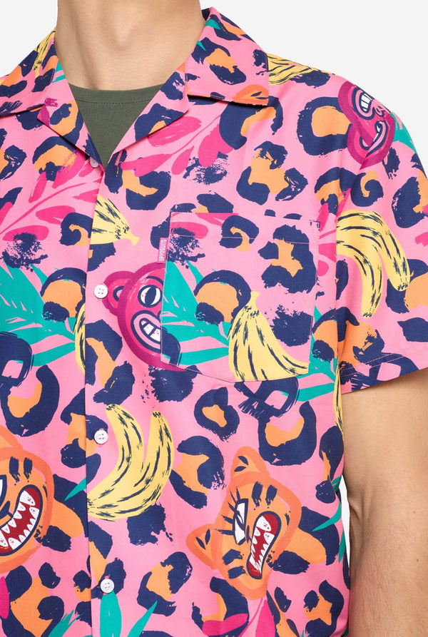 Ein Mann trägt ein farbenfrohes zweiteiliges Sommeroutfit mit auffälligem Tropenmuster. Das kurzärmelige Hemd und die dazu passenden Shorts präsentieren ein lebendiges Design mit neonpinken, orangefarbenen und gelben Leopardenmustern, Bananen, grünen Palmblättern und skurrilen Cartoon-Gesichtern. Das Outfit ist lässig mit einem grünen T-Shirt darunter gestylt und perfekt für Sommerpartys, Urlaube oder Spaß am Pool. Brust-Nahaufnahme.