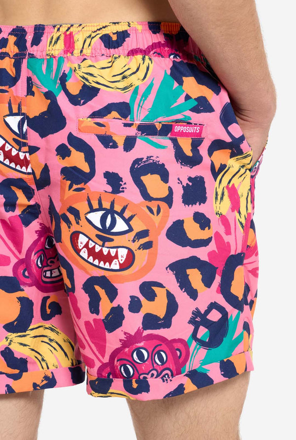 Ein Mann trägt ein farbenfrohes zweiteiliges Sommeroutfit mit auffälligem Tropenmuster. Das kurzärmelige Hemd und die dazu passenden Shorts präsentieren ein lebendiges Design mit neonpinken, orangefarbenen und gelben Leopardenmustern, Bananen, grünen Palmblättern und skurrilen Cartoon-Gesichtern. Das Outfit ist lässig mit einem grünen T-Shirt darunter gestylt und perfekt für Sommerpartys, Urlaube oder Spaß am Pool. Nahaufnahme der Rückseite der Shorts.