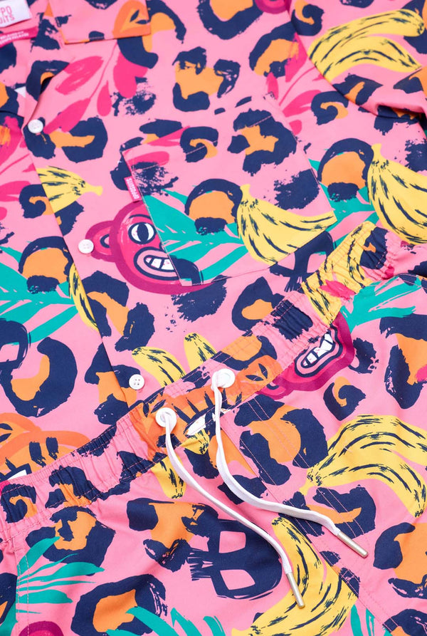 Bild eines farbenfrohen zweiteiligen Sommeroutfits mit auffälligem Tropenmuster. Das kurzärmelige Hemd und die dazu passenden Shorts präsentieren ein lebendiges Design mit neonpinken, orangefarbenen und gelben Leopardenmustern, Bananen, grünen Palmblättern und witzigen Cartoon-Gesichtern. Das Outfit ist lässig mit einem grünen T-Shirt darunter gestylt und perfekt für Sommerpartys, Urlaube oder Spaß am Pool. Nahaufnahme.