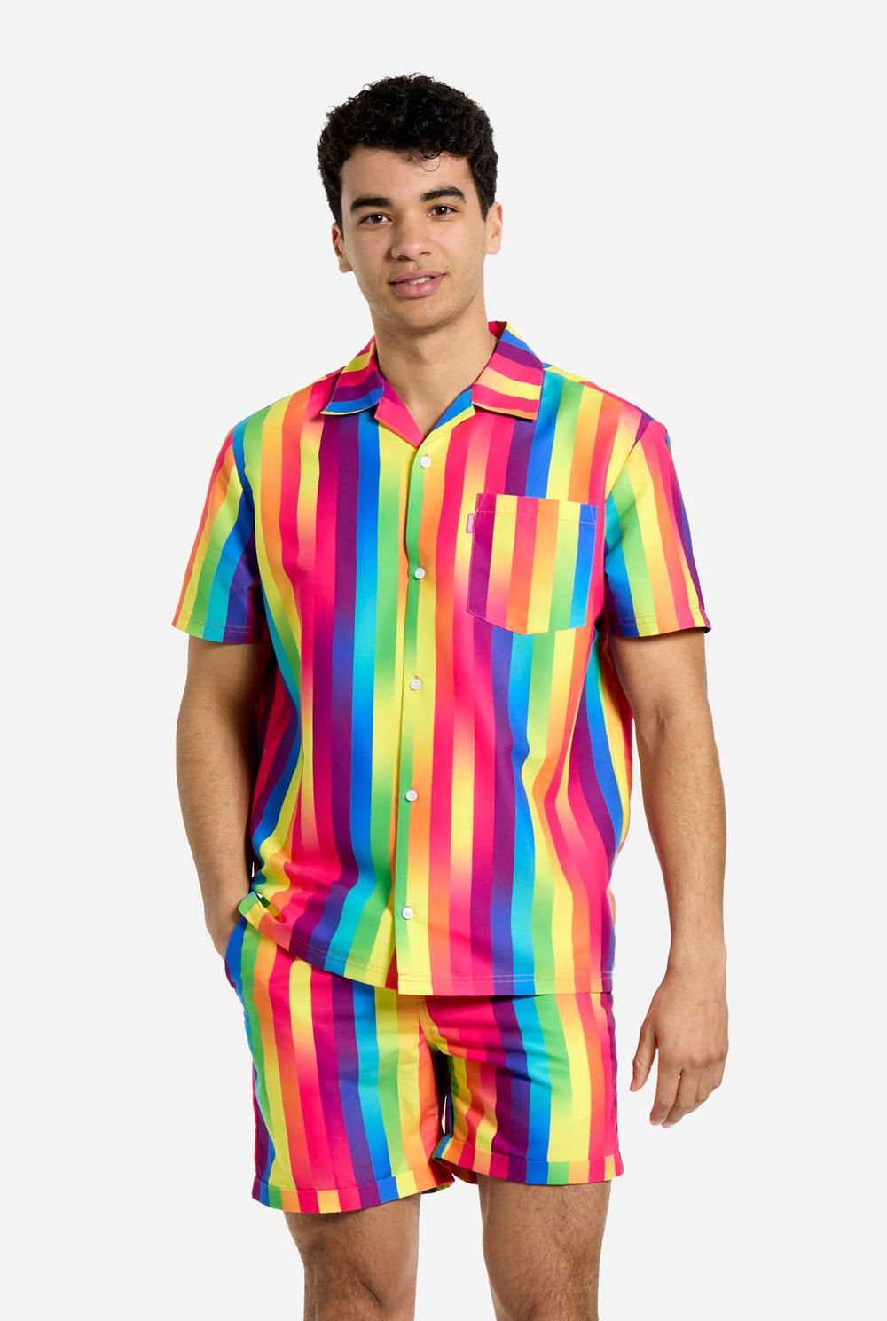 Ein Mann trägt ein vertikal gestreiftes, regenbogenfarbenes Sommerset, bestehend aus Hemd und Shorts. Er lächelt in die Kamera und hält eine Hand in der Tasche.
