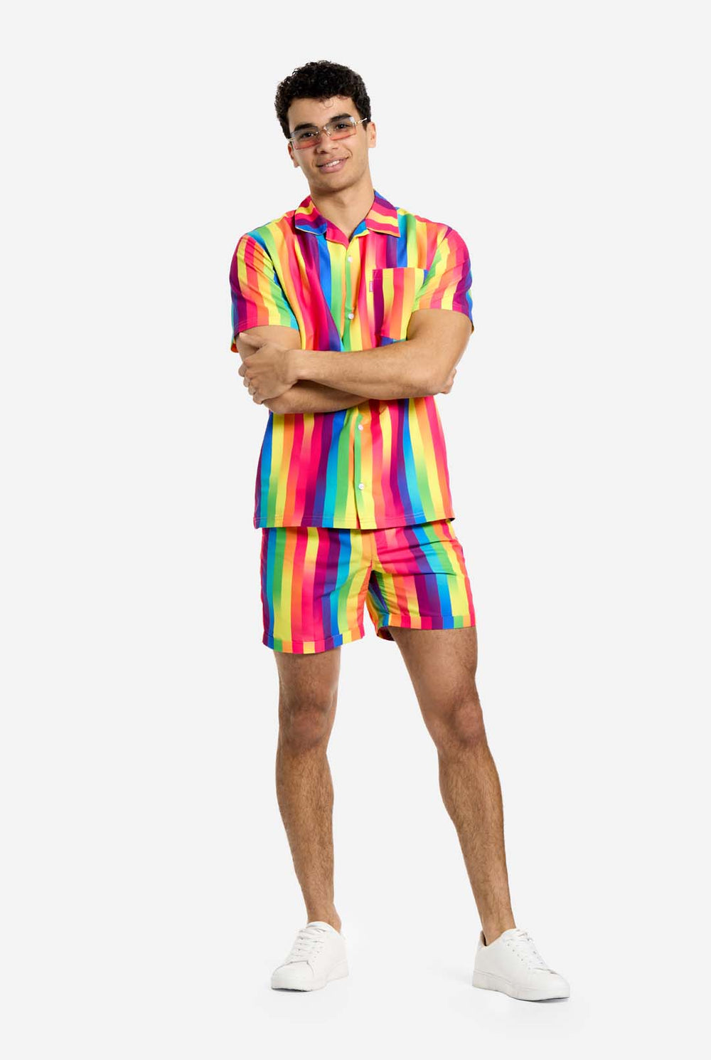 Ein Mann trägt ein vertikal gestreiftes, regenbogenfarbenes Sommerset, bestehend aus Hemd und Shorts. Er lächelt in die Kamera und hält eine Hand in der Tasche.
