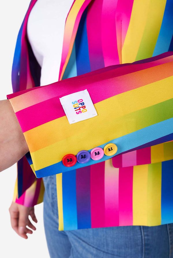 Nahaufnahme einer Frau in einem vertikal gestreiften Damenblazer in Regenbogenfarben, einem weißen Hemd und blauen Jeans. Sie zeigt den Ärmel mit farbigen Knöpfen.