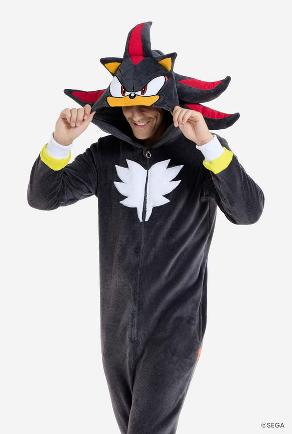 Ein Mann, der einen schwarzen Plüsch-Onesie trägt, der von der beliebten Igel-Figur Shadow inspiriert ist, mit stacheligen roten und schwarzen Akzenten an der Kapuze, einem weißen Brustemblem und gelben Details an den Handgelenken. Der Strampler ist aus weichem Fleecematerial gefertigt und bietet Komfort und ein verspieltes Design für Cosplay, Faulenzen oder thematische Veranstaltungen. Die Person lächelt, während sie die Kapuze zurechtrückt, und zeigt dabei das erkennbare Gesicht und das auffällige Design 