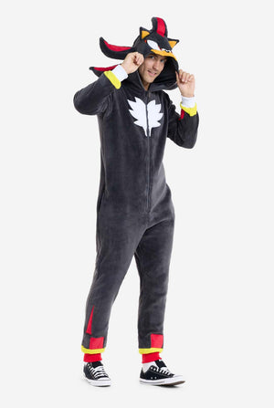 Ein Mann trägt einen plüschigen schwarzen Onesie, inspiriert vom beliebten Igel Shadow. Er besticht durch stachelige rote und schwarze Akzente an der Kapuze, ein weißes Brustemblem und gelbe Handgelenksdetails. Der Onesie aus weichem Fleece bietet Komfort und ein verspieltes Design für Cosplay, zum Entspannen oder für Themenveranstaltungen. Die Person lächelt, während sie die Kapuze hält und das bekannte Gesicht und das auffällige Design der Figur zeigt. Perfekt für Fans von Videospielen oder Zeichentrickfi