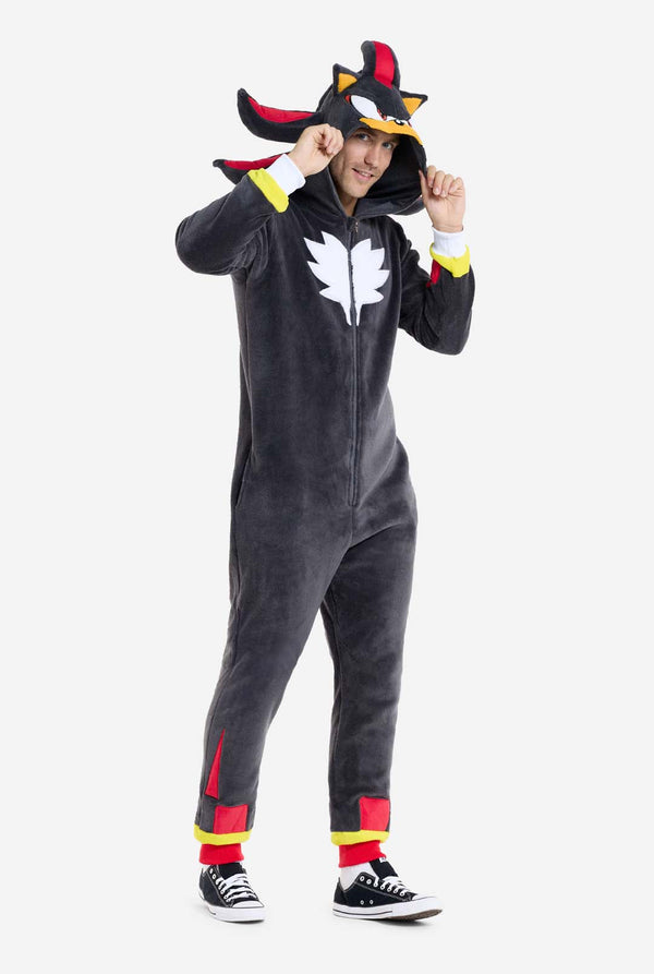 Ein Mann trägt einen plüschigen schwarzen Onesie, inspiriert vom beliebten Igel Shadow. Er besticht durch stachelige rote und schwarze Akzente an der Kapuze, ein weißes Brustemblem und gelbe Handgelenksdetails. Der Onesie aus weichem Fleece bietet Komfort und ein verspieltes Design für Cosplay, zum Entspannen oder für Themenveranstaltungen. Die Person lächelt, während sie die Kapuze hält und das bekannte Gesicht und das auffällige Design der Figur zeigt. Perfekt für Fans von Videospielen oder Zeichentrickfi