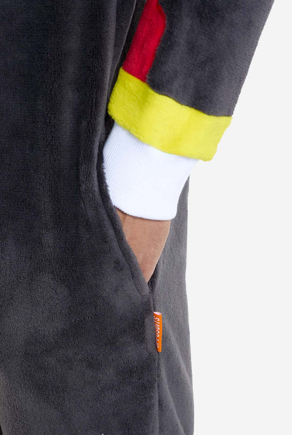 Ein Mann trägt einen plüschigen schwarzen Onesie, inspiriert vom beliebten Igel Shadow. Er besticht durch stachelige rote und schwarze Akzente an der Kapuze, ein weißes Brustemblem und gelbe Handgelenksdetails. Der Onesie aus weichem Fleece bietet Komfort und ein verspieltes Design für Cosplay, zum Entspannen oder für Themenveranstaltungen. Perfekt für Fans von Gaming- oder Zeichentrickfiguren. Ärmelnahaufnahme mit Hand in der Tasche.