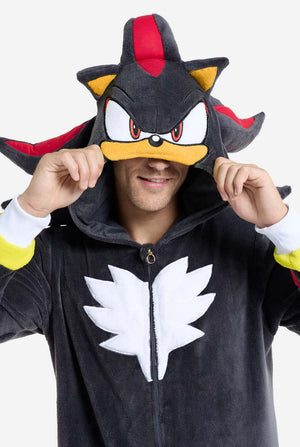 Ein Mann trägt einen plüschigen schwarzen Onesie, inspiriert vom beliebten Igel Shadow. Er besticht durch stachelige rote und schwarze Akzente an der Kapuze, ein weißes Brustemblem und gelbe Handgelenksdetails. Der Onesie aus weichem Fleece bietet Komfort und ein verspieltes Design für Cosplay, zum Entspannen oder für Themenveranstaltungen. Lächelnd rückt die Person die Kapuze zurecht und zeigt das bekannte Gesicht und das auffällige Design der Figur. Perfekt für Fans von Videospielen oder Zeichentrickfigur