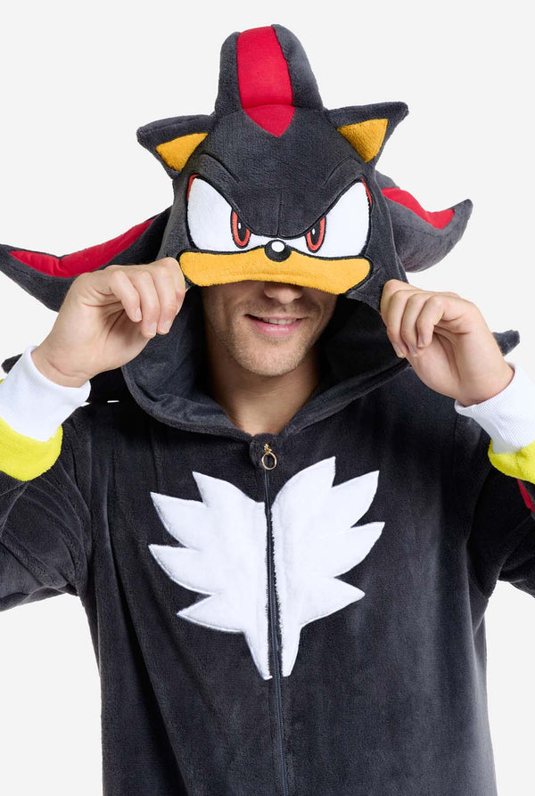 Ein Mann trägt einen plüschigen schwarzen Onesie, inspiriert vom beliebten Igel Shadow. Er besticht durch stachelige rote und schwarze Akzente an der Kapuze, ein weißes Brustemblem und gelbe Handgelenksdetails. Der Onesie aus weichem Fleece bietet Komfort und ein verspieltes Design für Cosplay, zum Entspannen oder für Themenveranstaltungen. Lächelnd rückt die Person die Kapuze zurecht und zeigt das bekannte Gesicht und das auffällige Design der Figur. Perfekt für Fans von Videospielen oder Zeichentrickfigur
