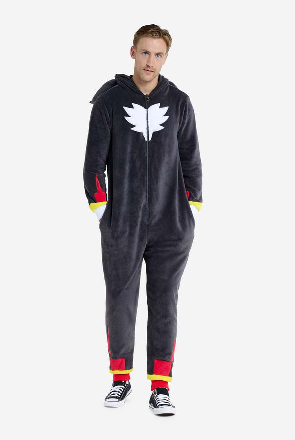 Ein Mann trägt einen plüschigen schwarzen Onesie, inspiriert vom beliebten Igel Shadow. Er besticht durch stachelige rote und schwarze Akzente an der Kapuze, ein weißes Brustemblem und gelbe Handgelenksdetails. Der Onesie aus weichem Fleece bietet Komfort und ein verspieltes Design für Cosplay, Lounge-Abende oder Themenveranstaltungen. Die Person lächelt, während die Kapuze auf ihrem Rücken liegt. Perfekt für Fans von Gaming- oder Zeichentrickfiguren. Ganzkörperbild.