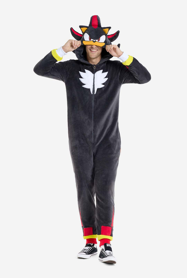 Ein Mann trägt einen plüschigen schwarzen Onesie, inspiriert vom beliebten Igel Shadow. Er besticht durch stachelige rote und schwarze Akzente an der Kapuze, ein weißes Brustemblem und gelbe Handgelenksdetails. Der Onesie aus weichem Fleece bietet Komfort und ein verspieltes Design für Cosplay, Lounge-Abende oder Themenveranstaltungen. Lächelnd rückt die Person die Kapuze zurecht und zeigt das bekannte Gesicht und das auffällige Design der Figur. Perfekt für Fans von Videospielen oder Zeichentrickfiguren. 