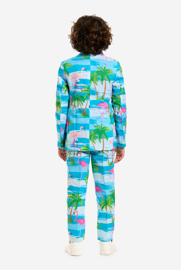 Auf diesem Bild trägt ein Kind einen hellblauen Jungenanzug mit tropischem Aufdruck und ein weißes Hemd. Der Anzug hat einen blauen Aufdruck, der an Wasser, rosa Flamingos und grüne Palmen erinnert.