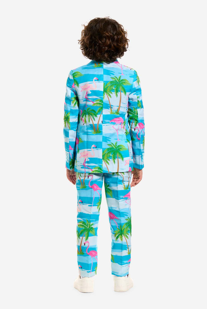 Auf diesem Bild trägt ein Kind einen hellblauen Jungenanzug mit tropischem Aufdruck und ein weißes Hemd. Der Anzug hat einen blauen Aufdruck, der an Wasser, rosa Flamingos und grüne Palmen erinnert.