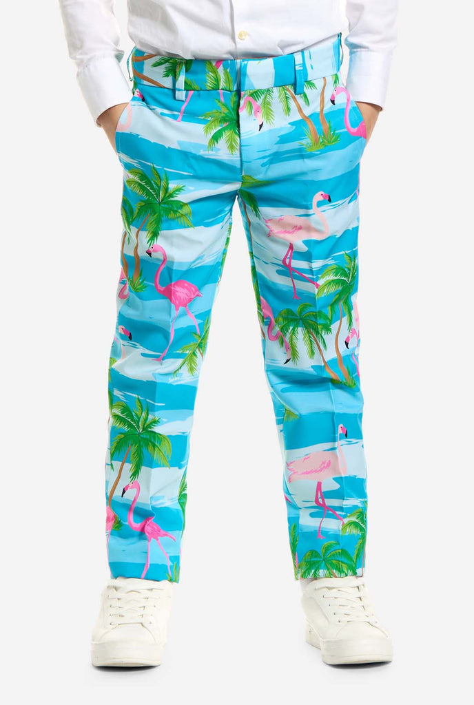 Auf diesem Bild trägt ein Kind einen hellblauen Jungenanzug mit tropischem Aufdruck und ein weißes Hemd. Der Anzug hat einen blauen Aufdruck, der an Wasser, rosa Flamingos und grüne Palmen erinnert.