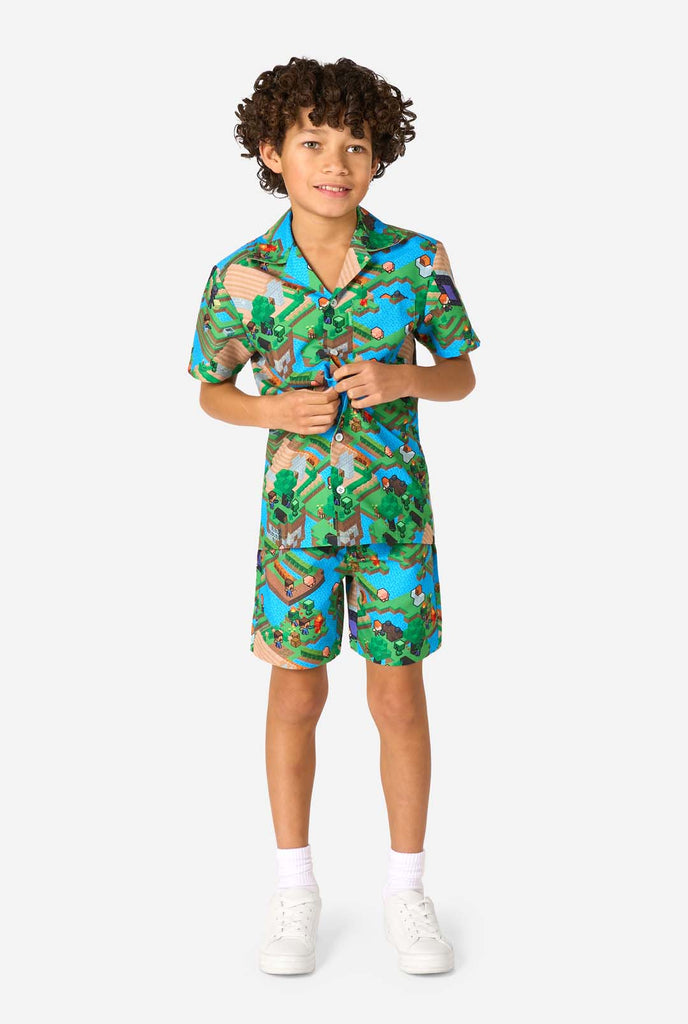 Ein Junge trägt ein buntes Sommeroutfit mit einem Minecraft-Overworld-Motiv, das pixelige Landschaften, Charaktere und Elemente aus dem ikonischen Spiel zeigt. Das kurzärmelige Hemd und die passenden Shorts heben ein lebhaftes Design hervor, das von der Minecraft-Welt inspiriert ist. Ganzkörperbild.