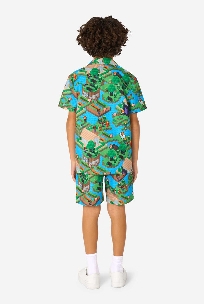Ein Junge trägt ein buntes Sommeroutfit mit einem Minecraft-Overworld-Motiv, das pixelige Landschaften, Charaktere und Elemente aus dem ikonischen Spiel zeigt. Das kurzärmelige Hemd und die passenden Shorts heben ein lebhaftes Design hervor, das von der Minecraft-Welt inspiriert ist. Ansicht von hinten.