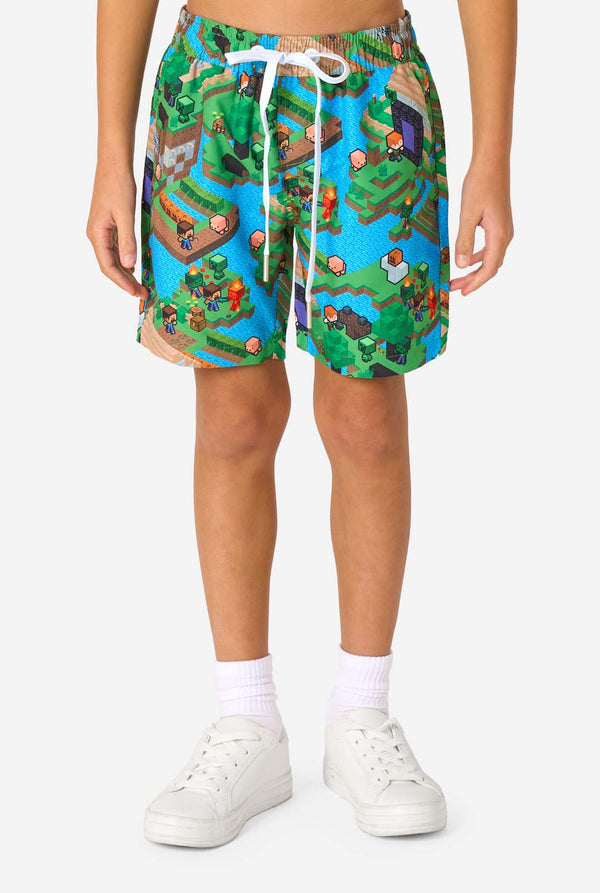 Ein Junge trägt ein buntes Sommeroutfit mit einem Minecraft-Overworld-Motiv, das pixelige Landschaften, Charaktere und Elemente aus dem ikonischen Spiel zeigt. Das kurzärmelige Hemd und die passenden Shorts heben ein lebhaftes Design hervor, das von der Minecraft-Welt inspiriert ist. Nahaufnahme der Shorts.