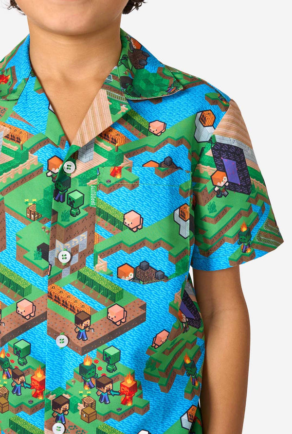 Ein Junge trägt ein buntes Sommeroutfit mit einem Minecraft-Overworld-Motiv, das pixelige Landschaften, Charaktere und Elemente aus dem ikonischen Spiel zeigt. Das kurzärmelige Hemd und die passenden Shorts heben ein lebhaftes Design hervor, das von der Minecraft-Welt inspiriert ist. Nahaufnahme der Brust.