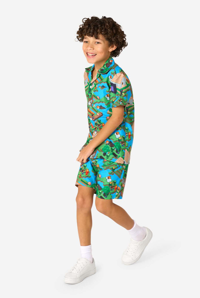 Ein Junge trägt ein buntes Sommeroutfit mit einem Minecraft-Overworld-Motiv, das pixelige Landschaften, Charaktere und Elemente aus dem ikonischen Spiel zeigt. Das kurzärmelige Hemd und die passenden Shorts heben ein lebhaftes Design hervor, das von der Minecraft-Welt inspiriert ist. Er geht.
