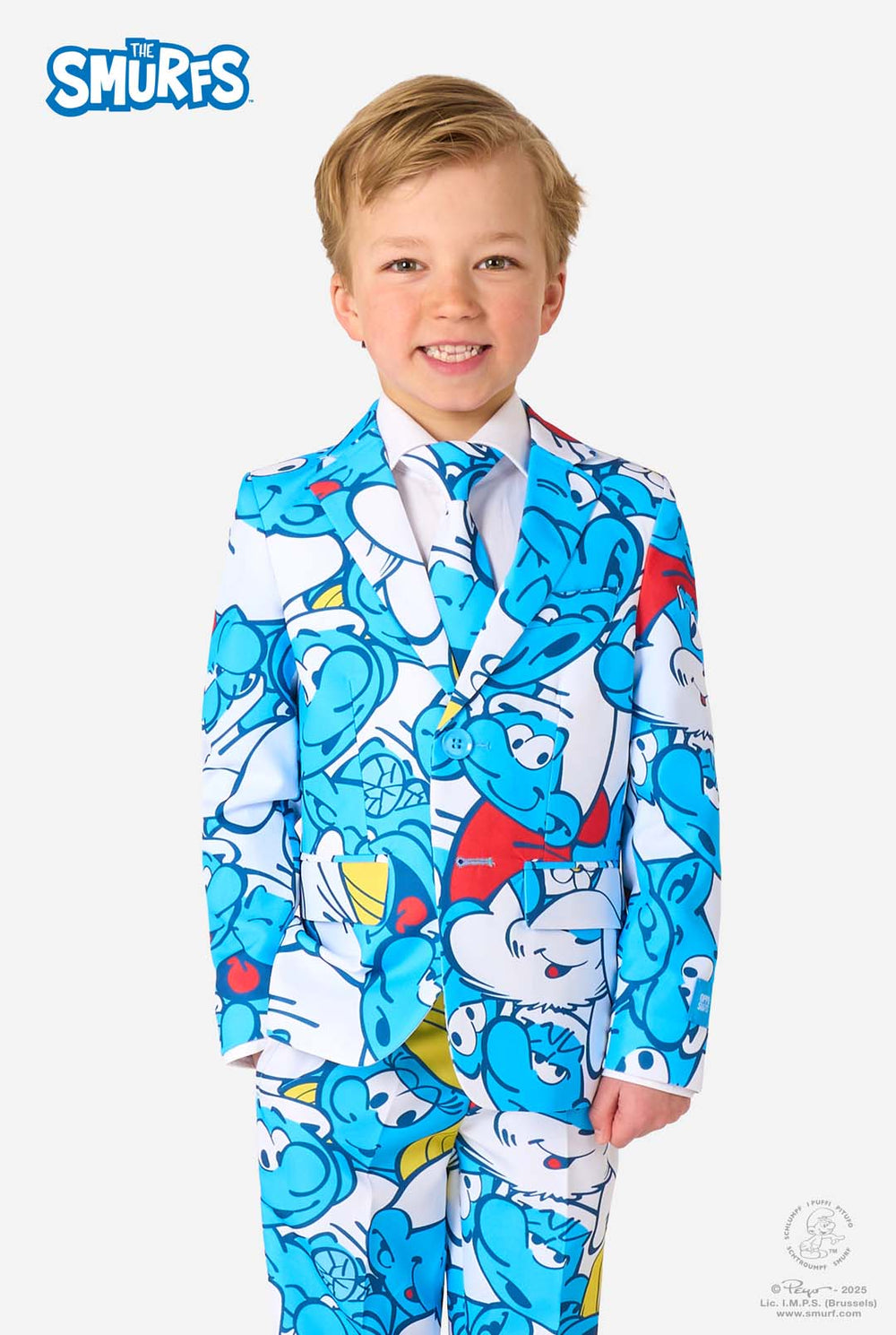 Junger Junge trägt einen bunten Kinderanzug mit Schlumpf-Motiv, der lebendige blaue Cartoon-Figuren zeigt, inklusive Jacke, Hose und passender Krawatte, lächelt vor einem schlichten weißen Hintergrund.