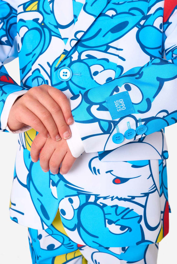 Junger Junge trägt einen farbenfrohen Schlümpfe-Kinderanzug mit lebendigen blauen Cartoon-Motiven, einschließlich Jacke, Hose und passender Krawatte, und lächelt vor einem schlichten weißen Hintergrund. Nahaufnahme des Ärmels.