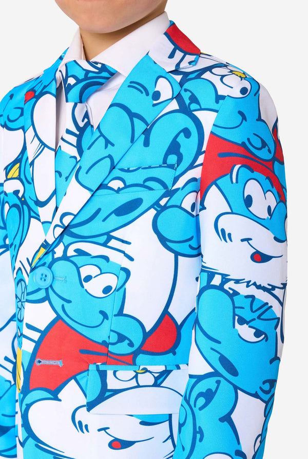 Junger Junge trägt einen farbenfrohen Kindersmoking mit Schlümpfe-Motiv und lebhaften blauen Cartoon-Prints, einschließlich Jacke, Hose und passender Krawatte, lächelt vor weißem Hintergrund. Nahaufnahme des Oberkörpers.