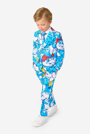 Junger Junge trägt einen farbenfrohen Kindersmoking mit Schlümpfe-Motiv und lebhaften blauen Cartoon-Prints, bestehend aus Jacke, Hose und passender Krawatte, lächelt vor weißem Hintergrund. Ganzkörperaufnahme, blickt auf den Boden.