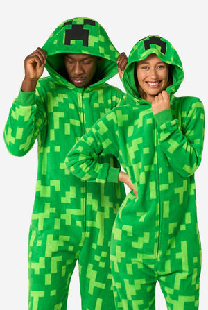 Zwei Personen tragen grüne Pixel-Strampler, die wie die Minecraft Creeper-Figur aussehen sollen. Die Onesies haben Kapuzen mit dem ikonischen schwarzen Pixelgesicht des Creepers. Der Mann hält seine Kapuze und blickt nach unten, während die Frauen mit einer Hand in der Hüfte in die Kamera lächeln