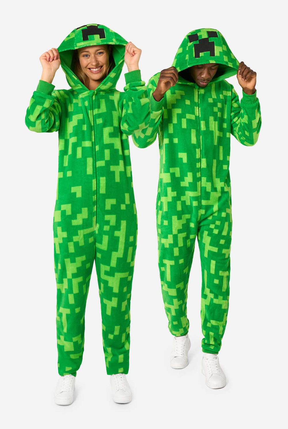 Zwei Personen tragen grüne Pixel-Strampler, die wie die Minecraft Creeper-Figur aussehen sollen. Die Onesies haben Kapuzen mit dem ikonischen schwarzen Pixelgesicht des Creepers. Der Mann hält seine Kapuze und blickt nach unten, während die Frauen mit einer Hand in der Hüfte in die Kamera lächeln