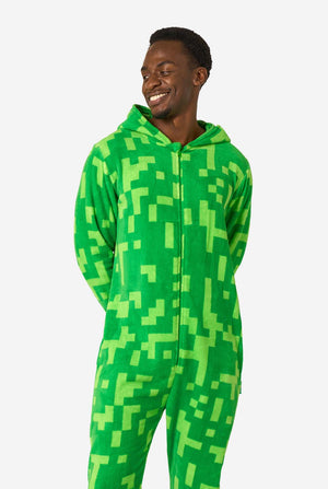 Ein Mann, der einen grünen, pixeligen Einteiler trägt, der wie der Minecraft Creeper-Charakter aussehen soll. Der Strampler hat eine Kapuze mit dem ikonischen schwarzen Pixelgesicht des Creepers. Der Mann hat die Kapuze auf dem Rücken und blickt zur Seite.
