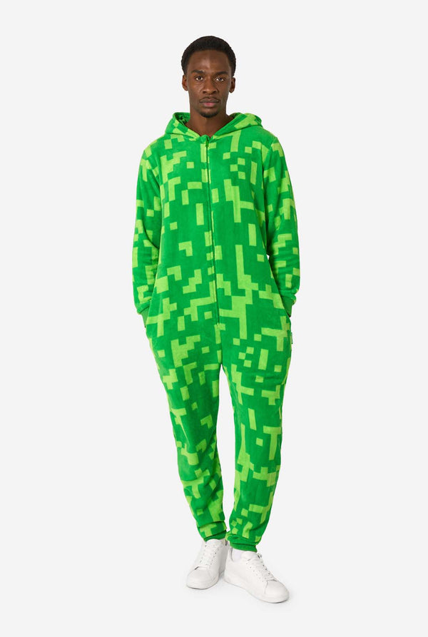 Ein Mann, der einen grünen, pixeligen Einteiler trägt, der wie der Minecraft Creeper-Charakter aussehen soll. Der Strampler hat eine Kapuze mit dem ikonischen schwarzen Pixelgesicht des Creepers. Der Mann hat die Kapuze auf dem Rücken und blickt in die Kamera, Ganzkörperaufnahme.