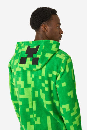 Ein Mann, der einen grünen, pixeligen Einteiler trägt, der wie der Minecraft Creeper-Charakter aussehen soll. Der Strampler hat eine Kapuze mit dem ikonischen schwarzen Pixelgesicht des Creepers. Der Mann schaut zur Seite. Ansicht von hinten.