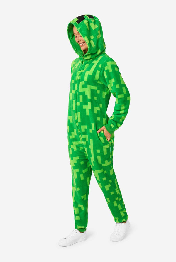 Eine Frau, die einen grünen, pixeligen Einteiler trägt, der wie die Minecraft Creeper-Figur aussehen soll. Der Strampler hat eine Kapuze mit dem ikonischen schwarzen Pixelgesicht des Creepers. Die Frau trägt die Kapuze und blickt auf den Boden.