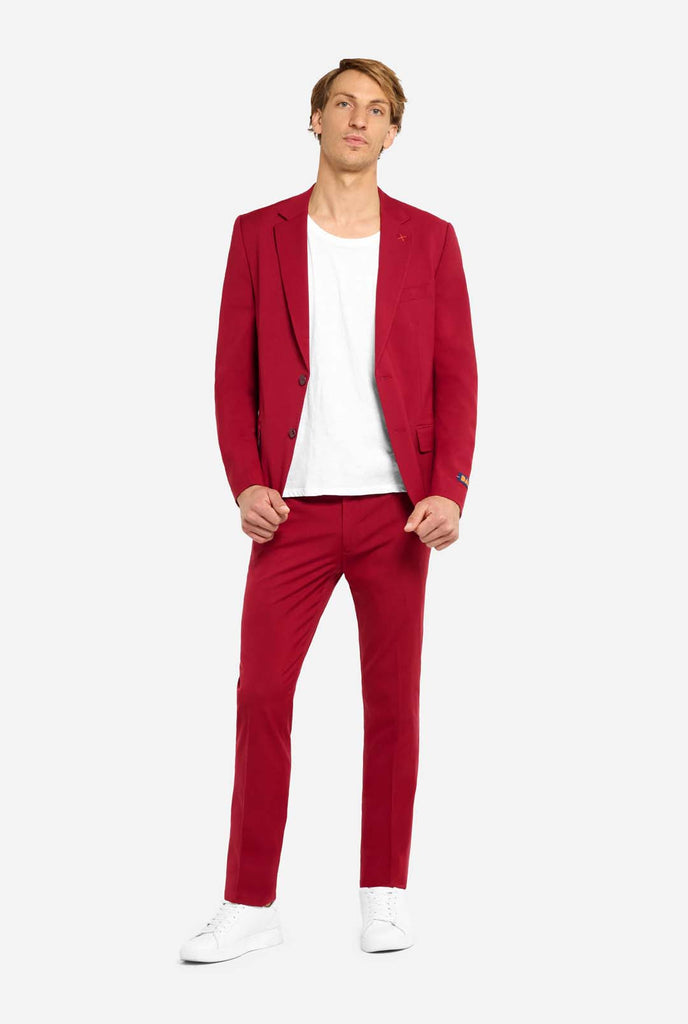 Bild mit hellgrauem Hintergrund und Mann trägt Bright Burgundy Herrenanzug und weißes Hemd. Anzüge besteht aus Blazer und Hose. Mann schaut in die Kamera, Ganzkörperbild.