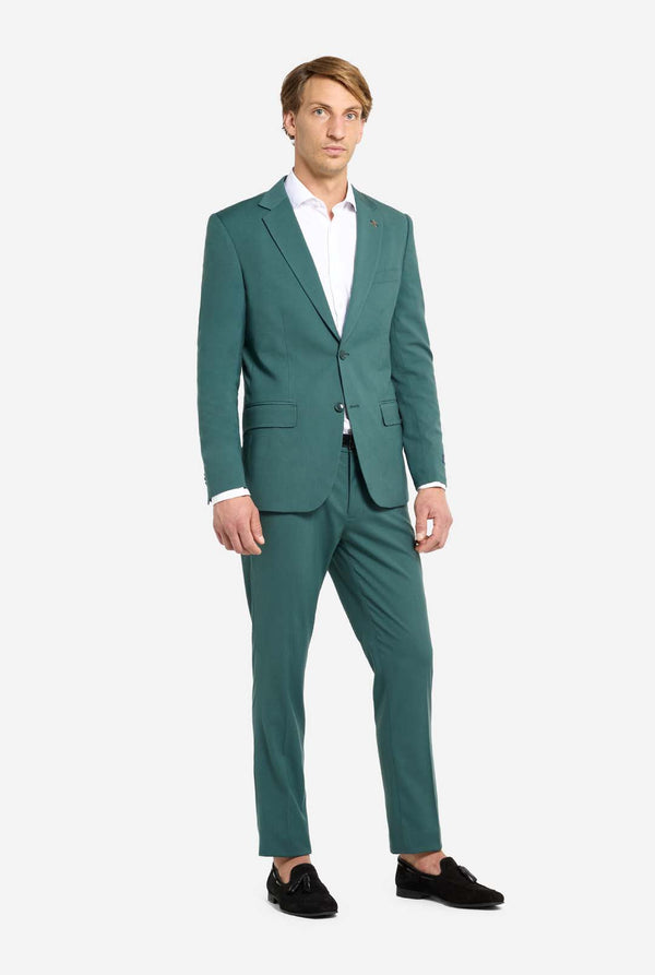 Bild mit hellgrauem Hintergrund und einem Mann in dunkelgrünem Anzug und weißem Hemd. Der Anzug besteht aus Blazer und Hose. Der Mann blickt in die Kamera. Ganzkörperaufnahme.