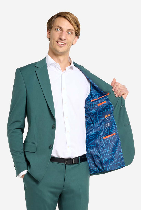 Bild mit hellgrauem Hintergrund und einem Mann in dunkelgrünem Herrenanzug und weißem Hemd. Der Anzug besteht aus Blazer und Hose. Der Mann blickt zur Seite und hält eine Hand in der Hosentasche, eine Seite der Jacke ist offen.