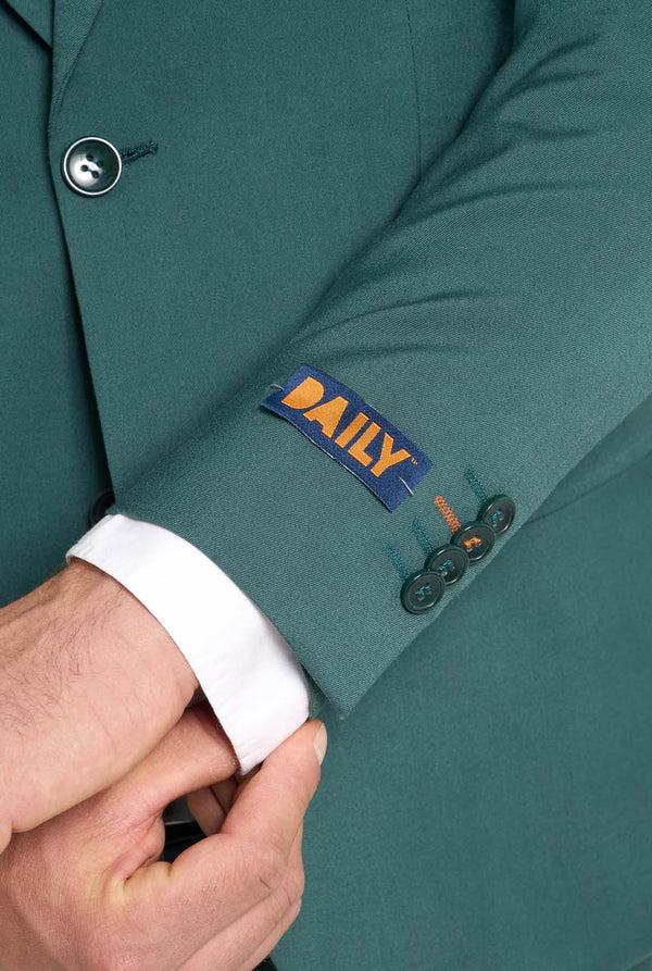 Bild mit hellgrauem Hintergrund und einem Mann in dunkelgrünem Anzug und weißem Hemd. Der Anzug besteht aus Blazer und Hose. Nahaufnahme der Ärmel.