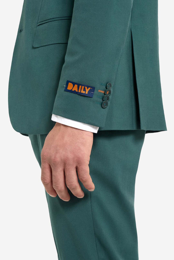 Bild mit hellgrauem Hintergrund und einem Mann in dunkelgrünem Anzug und weißem Hemd. Der Anzug besteht aus Blazer und Hose. Seitenansicht mit sichtbarem Hosenunterteil und Blazerunterteil.
