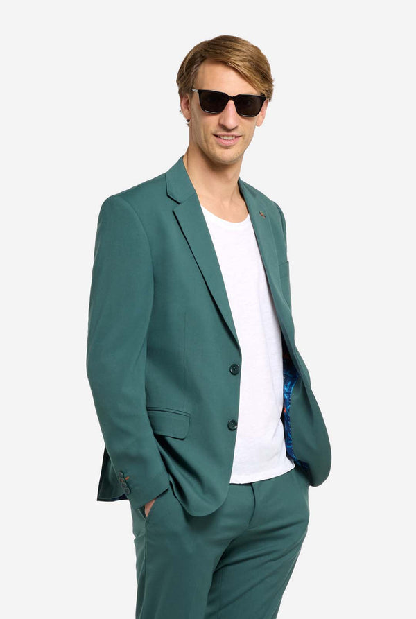 Bild mit hellgrauem Hintergrund. Mann trägt dunkelgrünen Anzug und weißes T-Shirt. Anzug besteht aus Blazer und Hose. Mann blickt in die Kamera und trägt eine Sonnenbrille.