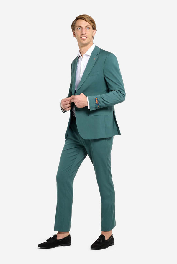 Bild mit hellgrauem Hintergrund und einem Mann in dunkelgrünem Anzug und weißem Hemd. Der Anzug besteht aus Blazer und Hose. Der Mann blickt zur Seite. Ganzkörperbild.